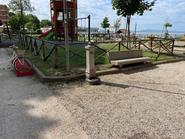 Terrain de jeu de Passignano. À gauche, un vélo blanc. Banc et fontaine à l'entrée de l'aire de jeux clôturée.